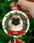 Personalized custom round ceramic Christmas ornament with a pug portrait in a festive wreath, "Bella" on a red banner.